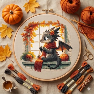 May include: A cross-stitch embroidery project in a wooden hoop featuring a small black dragon wearing a red scarf, sitting on a windowsill with autumn leaves and pumpkins. The design is surrounded by orange yarn, embroidery floss, and scissors.