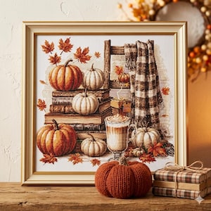 May include: A framed cross-stitch artwork depicts a fall scene with pumpkins, books, and a cozy blanket. The pumpkins are various sizes and colors, with orange and white hues. A latte sits next to the pumpkins. Autumn leaves are scattered throughout the scene.
