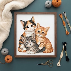 Puede incluir: Un bordado en punto de cruz enmarcado que muestra tres gatitos: un calicó, un atigrado naranja y un atigrado gris. La obra se presenta con hilo de bordar, ovillos de lana y tijeras. Los gatitos están representados sobre una tela blanca.