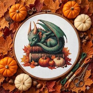 May include: A circular wooden embroidery hoop containing a cross-stitch pattern of a green dragon sleeping on a stack of books surrounded by autumn leaves and pumpkins. The design features orange, green, and white pumpkins with fall foliage.