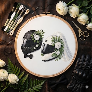 Op de afbeelding: Kruissteekborduurwerk in een houten ring met twee motorhelmen, één zwart en één wit, versierd met bloemen. Een zwarte vlinderdas bevindt zich onder de zwarte helm. Witte rozen, borduurgaren en een leren handschoen omringen de scène.
