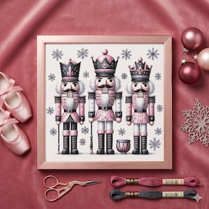 May include: A framed cross-stitch featuring three pink and black nutcracker figures with snowflake accents. The image includes ballet shoes, embroidery floss, and a pair of gold scissors, all set against a pink velvet backdrop.