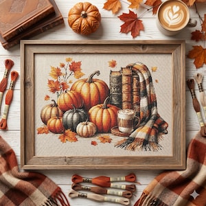 Puede incluir: Un bordado enmarcado con calabazas, libros y una bufanda. La obra presenta calabazas naranjas y grises, hojas de otoño y una taza de café. El marco de madera complementa la escena, creando un ambiente cálido y otoñal.