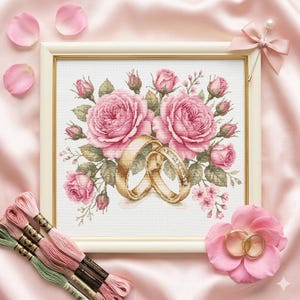 Op de afbeelding: Kruissteekkunstwerk met twee gouden trouwringen tegen een achtergrond van roze rozen en groen. Het ingelijste stuk is omgeven door roze rozenblaadjes, borduurgaren en een decoratieve strik, wat een romantisch thema creëert.