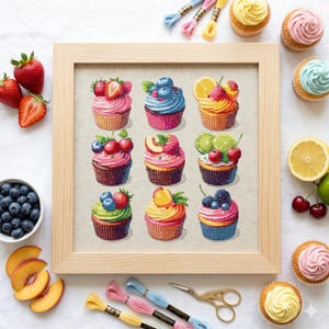 Op de afbeelding: Een ingelijste kruissteek met negen kleurrijke cupcakes, elk belegd met verschillende soorten fruit zoals aardbeien, blauwe bessen en perziken. De cupcakes staan in een houten lijst, met borduurgaren en een schaar. Vers fruit en cupcakes zijn rondom het frame geplaatst.
