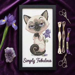 May include: A framed cross-stitch embroidery featuring a Siamese cat with purple eyes and a dark tail, sitting next to a purple iris flower. The text "Simply Fabulous" is stitched in black at the bottom of the white fabric within a black frame.