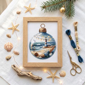 Peut inclure: Une broderie encadrée représentant un phare à l'intérieur d'une boule bleue, entourée de coquillages, d'étoiles de mer et de fils à broder. Le cadre en bois est rectangulaire. Une branche de conifère et des guirlandes lumineuses complètent le thème de Noël côtier.