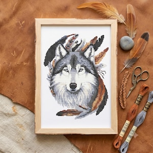 Pode incluir: Um bordado em ponto cruz de uma cabeça de lobo centralizada numa moldura de madeira clara. O lobo apresenta tons de pelo cinzento, branco e preto com penas castanhas e pretas atrás. O desenho é bordado em tecido branco com grelha visível.