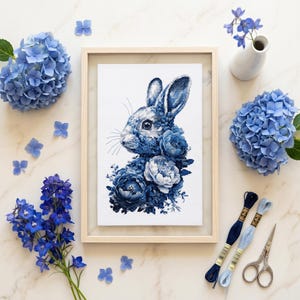 May include: A framed cross-stitch embroidery featuring a blue rabbit surrounded by blue floral patterns. The artwork is displayed with blue hydrangea blossoms, blue delphinium stems, two skeins of blue embroidery floss, and metal scissors.