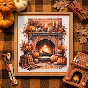 May include: A framed cross-stitch depicting a cozy fireplace scene with pumpkins, candles, and autumn foliage. The artwork features a brick fireplace with a roaring fire, surrounded by orange pumpkins and fall decorations. The frame is a light wood color.