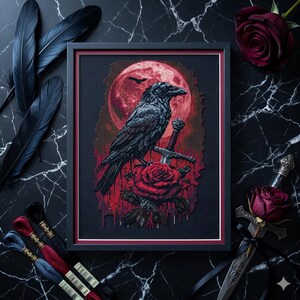 May include: Framed cross-stitch art featuring a black raven perched on a sword and red rose, set against a red moon backdrop. The artwork is in a black frame with a red border. Black feathers and embroidery floss are arranged around the frame.