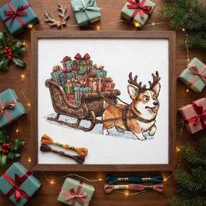 Op de afbeelding: Een ingelijst kruissteek borduurwerk met een Corgi die een rendiergewei draagt en een houten slee vol ingepakte kerstcadeaus trekt. Het kunstwerk ligt op een houten ondergrond, omringd door cadeauverpakkingen en dennentakken.