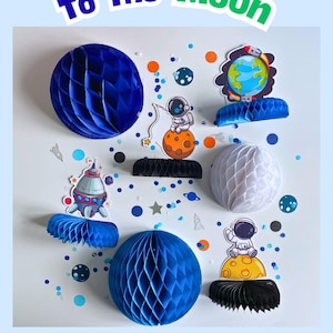 Peut inclure: Décorations sur le thème de l'espace, comprenant des boules en nid d'abeille bleues et blanches, des découpes d'astronautes et des motifs de fusées spatiales. Le texte "To The Moon" et "SPACE IN BOX" est affiché.