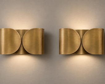 May include: A pair of wall sconces featuring a curved, metallic brass finish with two overlapping circular plates. The design emits a soft, warm glow from the top and bottom edges, creating a focused light effect against the wall surface.