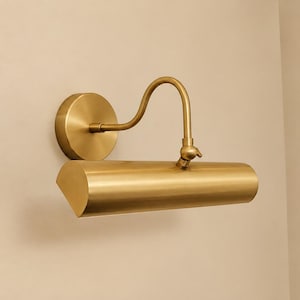 May include: A wall-mounted picture light with a brushed brass finish. The fixture features a circular wall plate, a curved arm, and a cylindrical, semi-circular shade that directs light downward. The design is metallic with a smooth, uniform texture.