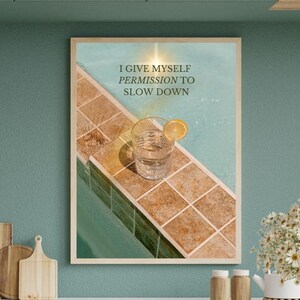 Könnte beinhalten: Ein gerahmter Druck mit dem Text "I GIVE MYSELF PERMISSION TO SLOW DOWN" zeigt ein Glas Wasser mit einer Zitronenscheibe auf einer gefliesten Oberfläche neben einem Pool. Das Bild ist in einer Küche mit einer türkisfarbenen Wand und einer weißen Arbeitsplatte.