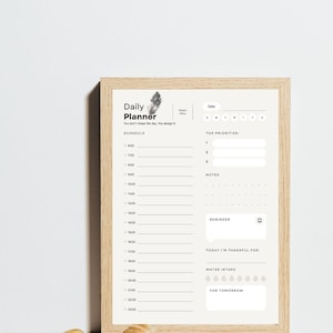 Może przedstawiać: Oprawiony planer dzienny z jasnobeżowym tłem i drewnianą ramą. Planer zawiera sekcje do planowania, najważniejsze priorytety, notatki, przypomnienia i spożycie wody. Tekst "Daily Planner" znajduje się na górze.