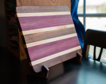 May include: A rectangular wooden cutting board with a striped pattern of purple, brown, and white wood. The board is standing on a black surface.