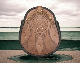 May include: A brown leather sporran with a decorative Celtic knot design and gold-toned studs. The sporran has a fringe and is hanging against a backdrop of water and sky.