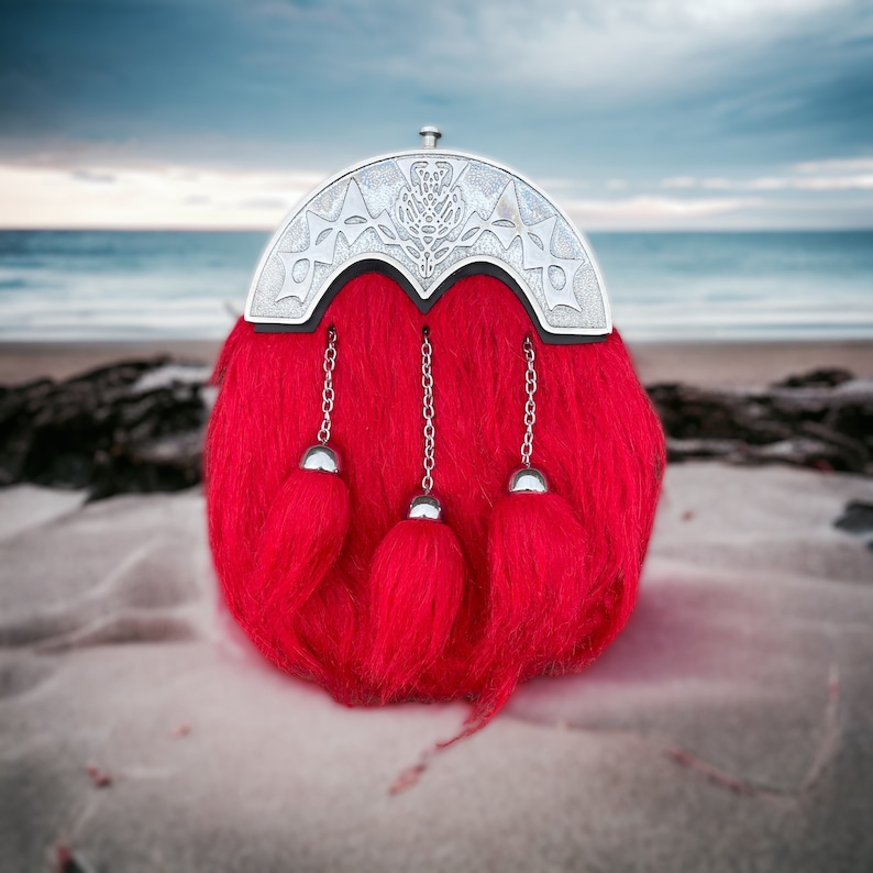 May include: A red kilt sporran with silver accents and a thistle design. The sporran has three tassels and is made of a soft, fuzzy material.