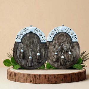 Puede incluir: Dos sporrans escoceses tradicionales con textura de piel, detalles plateados y borlas decorativas. Se presentan sobre una base de madera con vegetación, sobre un fondo neutro. Un accesorio clásico.