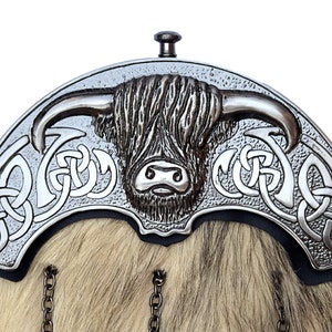 Op de afbeelding: Een zilveren kiltpin met een Hooglandkoe-ontwerp en Keltische knoopwerk. De pin is bevestigd aan een ketting.
