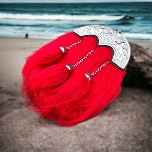 May include: A red kilt pin with a silver metal design. The pin has three tassels and is attached to a red kilt.