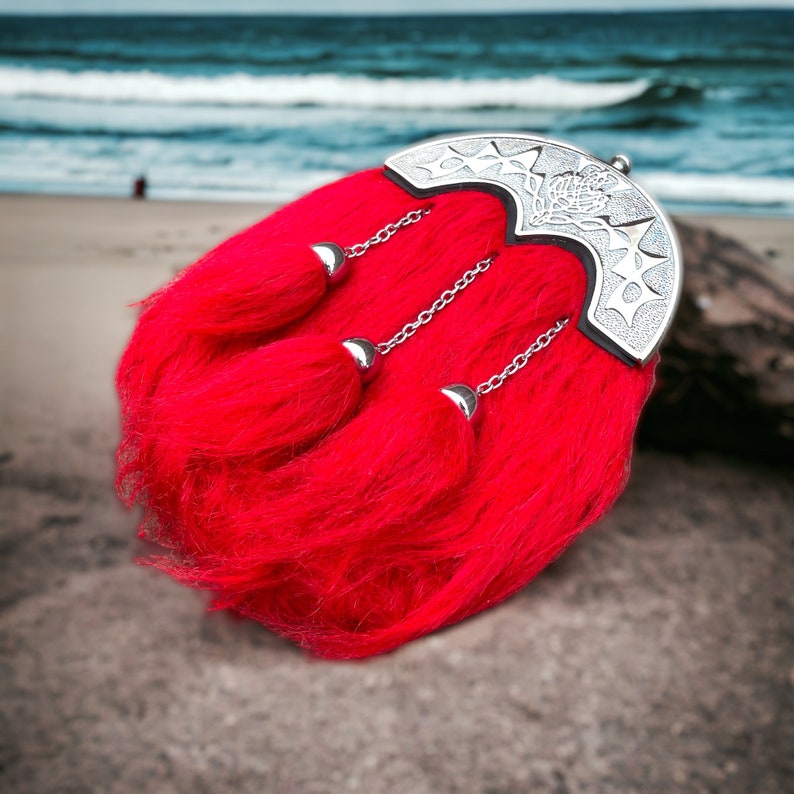 May include: A red kilt pin with a silver metal design. The pin has three tassels and is attached to a red kilt.