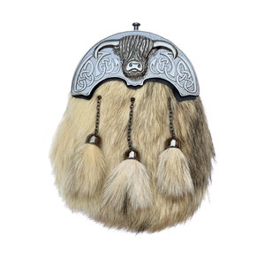 Op de afbeelding: Een zilveren kiltpin met een Hooglandkoe-ontwerp en Keltische knoopwerk. De pin is bevestigd aan een sporran van bruin en wit bont met drie kwastjes.
