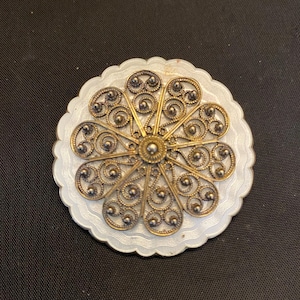 May include: A vintage-style brooch with a floral design. The brooch features a white, scalloped base with a filigree pattern in gold and silver tones. The central element is a gold-colored circle.