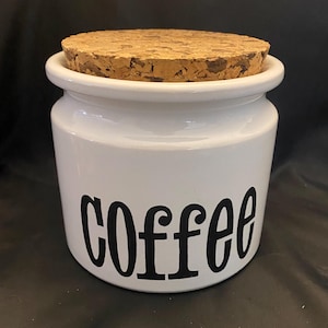 May include: White ceramic coffee canister with a cork lid. The word "coffee" is printed in black, bold letters on the front of the canister. The canister is cylindrical and has a rounded top.