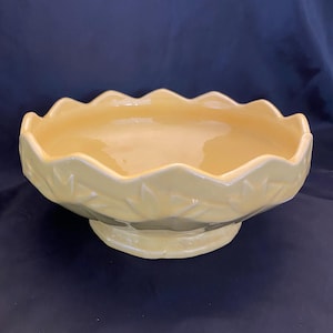 May include: A pale yellow ceramic bowl with a scalloped rim and a pedestal base. The bowl has a textured leaf pattern around the exterior. The interior of the bowl is smooth and glossy. The bowl is likely for decorative or serving purposes.