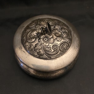 May include: A round, silver-toned container with a lid. The lid features an ornate floral and scroll design, with a small handle on top. The container appears to be made of metal and has a slightly aged appearance.