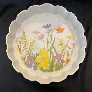 May include: White ceramic pie dish with a scalloped edge and a floral design. The design features colorful flowers including yellow, purple, pink, and orange blooms with green stems and leaves. The dish is round.