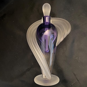May include: A decorative perfume bottle with a purple glass body and a frosted glass stopper. The bottle is supported by a clear, frosted glass base and features a swirling, wing-like design. The overall design is elegant and artistic.