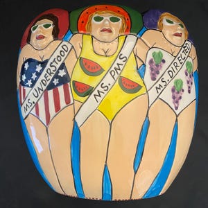 May include: A ceramic art piece featuring three figures in swimsuits. The figures wear sunglasses and sashes that read "MS. UNDERSTOOD", "MS. PMS", and "MS. DIRECTED". The swimsuits are decorated with stars and stripes, watermelon slices, and grapes.