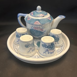 May include: A miniature ceramic tea set consisting of a small teapot with a lid and four matching cups arranged on a circular tray. The white porcelain features hand-painted blue and pink floral motifs, including lotus flowers and leaves.