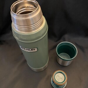 May include: An open, green Stanley thermos with a silver rim and a matching green and silver lid and cup. The Stanley logo is visible on the side of the thermos. The thermos is designed to keep liquids hot or cold.