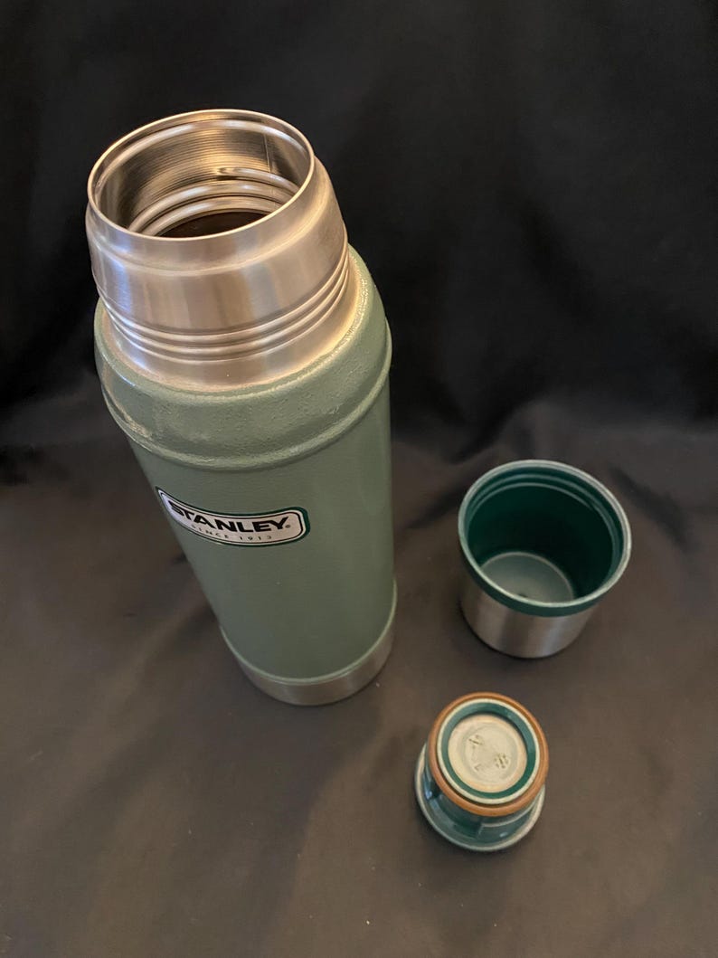May include: An open, green Stanley thermos with a silver rim and a matching green and silver lid and cup. The Stanley logo is visible on the side of the thermos. The thermos is designed to keep liquids hot or cold.
