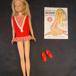 May include: A blonde Skipper doll wearing a red and white striped sleeveless top with a red bodice and matching shorts, accompanied by a pair of red shoes and a paper booklet titled Junior Edition Styles for Skipper Barbie's Little Sister.
