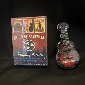May include: A deck of Spirit of Nashville playing cards in a rectangular box featuring illustrations and a circular logo, alongside a guitar-shaped metal tin with a red and black finish and the word Nashville printed on the front of the body.