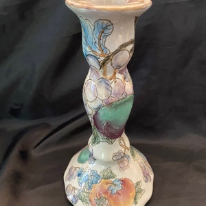 May include: A ceramic candlestick holder with a white base and a flared top. The candlestick is decorated with colorful illustrations of fruit, including grapes, apples, and plums, with green leaves and blue accents.