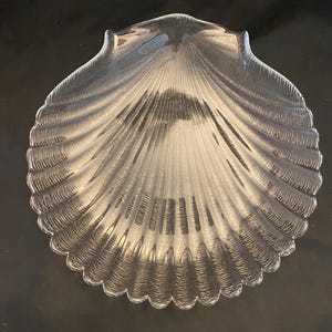 May include: Clear glass seashell-shaped dish. The dish has a scalloped edge and a textured surface with radiating lines, resembling the natural form of a seashell. The dish is on a black surface.