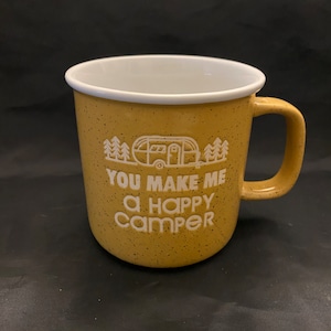 May include: Mustard yellow ceramic mug with a white rim and handle. The mug features the text "YOU MAKE ME A HAPPY CAMPER" in white, along with a graphic of a camper and trees. The mug has a speckled design.