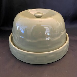 May include: A sage green ceramic cheese dome with a matching base. The dome has a rounded top with a small knob handle. The base is a shallow, circular dish with a textured rim. The set is designed for serving cheese.