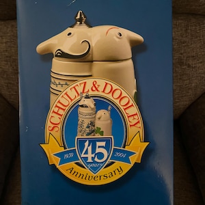 May include: A blue box with a white and red beer stein on the front. The stein is decorated with a white elephant and a green shamrock. The box has the text "Schlitz & Dooley" and "45 years Anniversary" on the front. The box also has the website "www.brewerysteins.com" on the bottom.