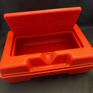 May include: A bright red, rectangular storage container with a textured surface. The lid is open, revealing an empty interior. The container has a boxy shape with rounded corners and a small, rectangular indentation on the front.