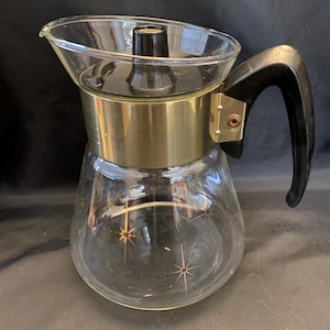 May include: A vintage glass coffee pot with a gold-colored metal band and a black handle. The clear glass carafe features starburst designs. The coffee pot has a black lid and a spout for pouring.