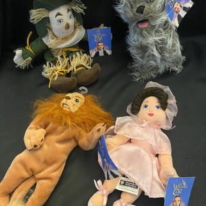 May include: Four plush dolls from The Wizard of Oz. The dolls include the Scarecrow, a gray dog, the Cowardly Lion, and Dorothy in a pink dress and ballet slippers. Each doll has a tag with the movie's logo.