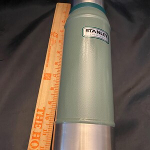 May include: A green Stanley thermos with a silver base and top. The thermos has a label that reads "STANLEY SINCE 1913". A wooden ruler is placed next to the thermos.